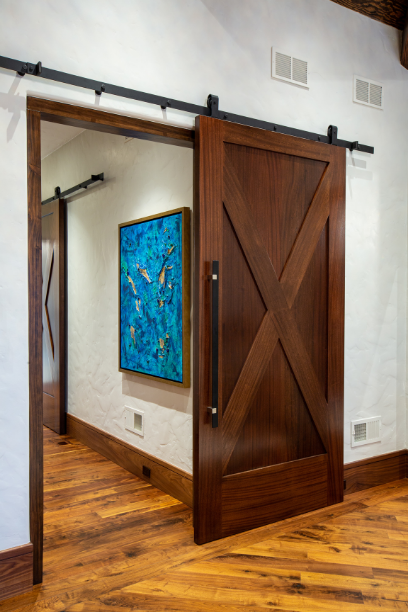 A wooden sliding barn door on metal rails opens to a hallway with hardwood floors, white textured walls, and a vibrant blue abstract painting hanging on the wall.