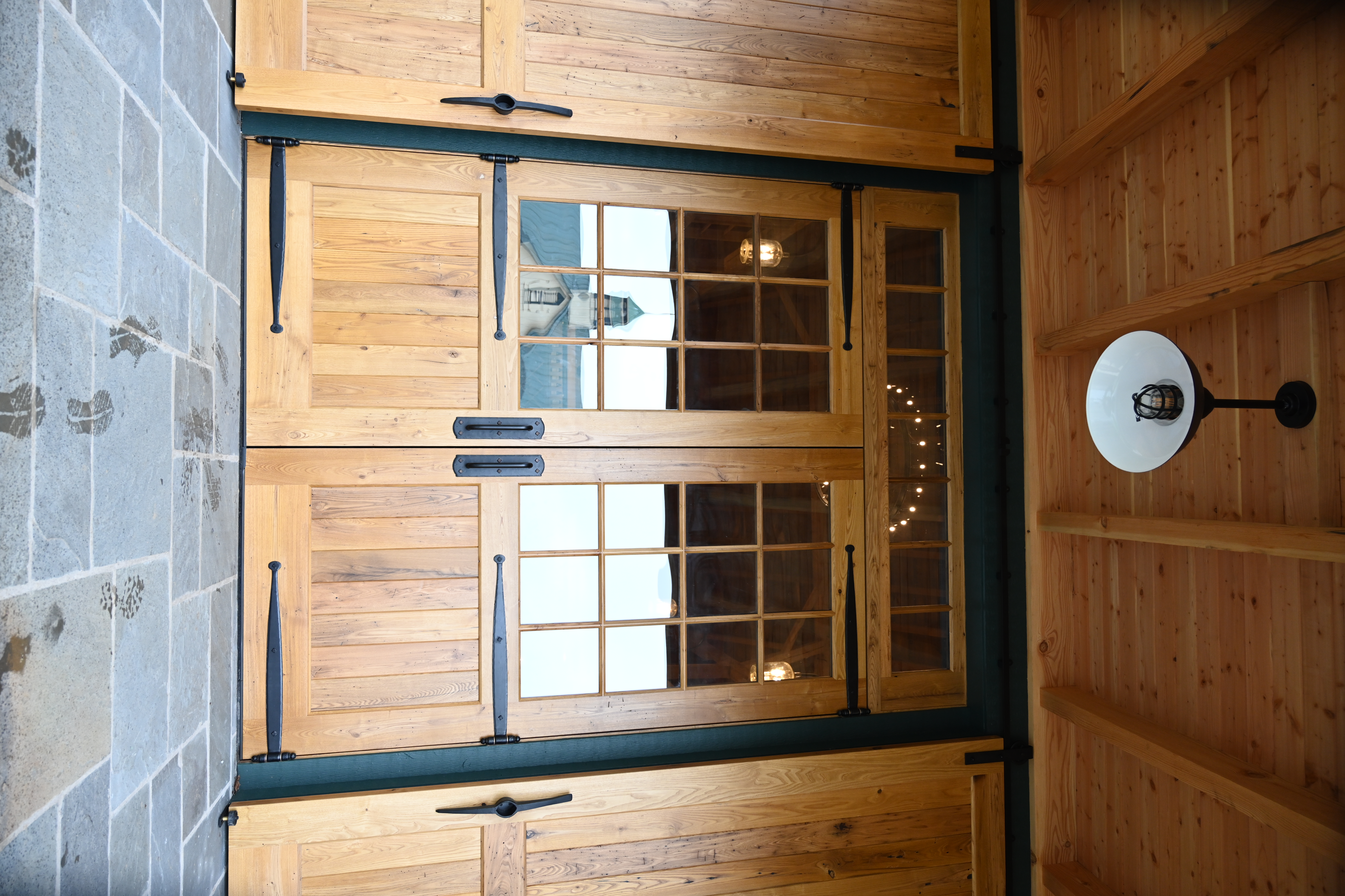 A wooden sliding barn door, partially open, reveals a bedroom with white bunk beds. The surrounding walls are made of horizontal light wood planks, and exposed beams run across the ceiling.