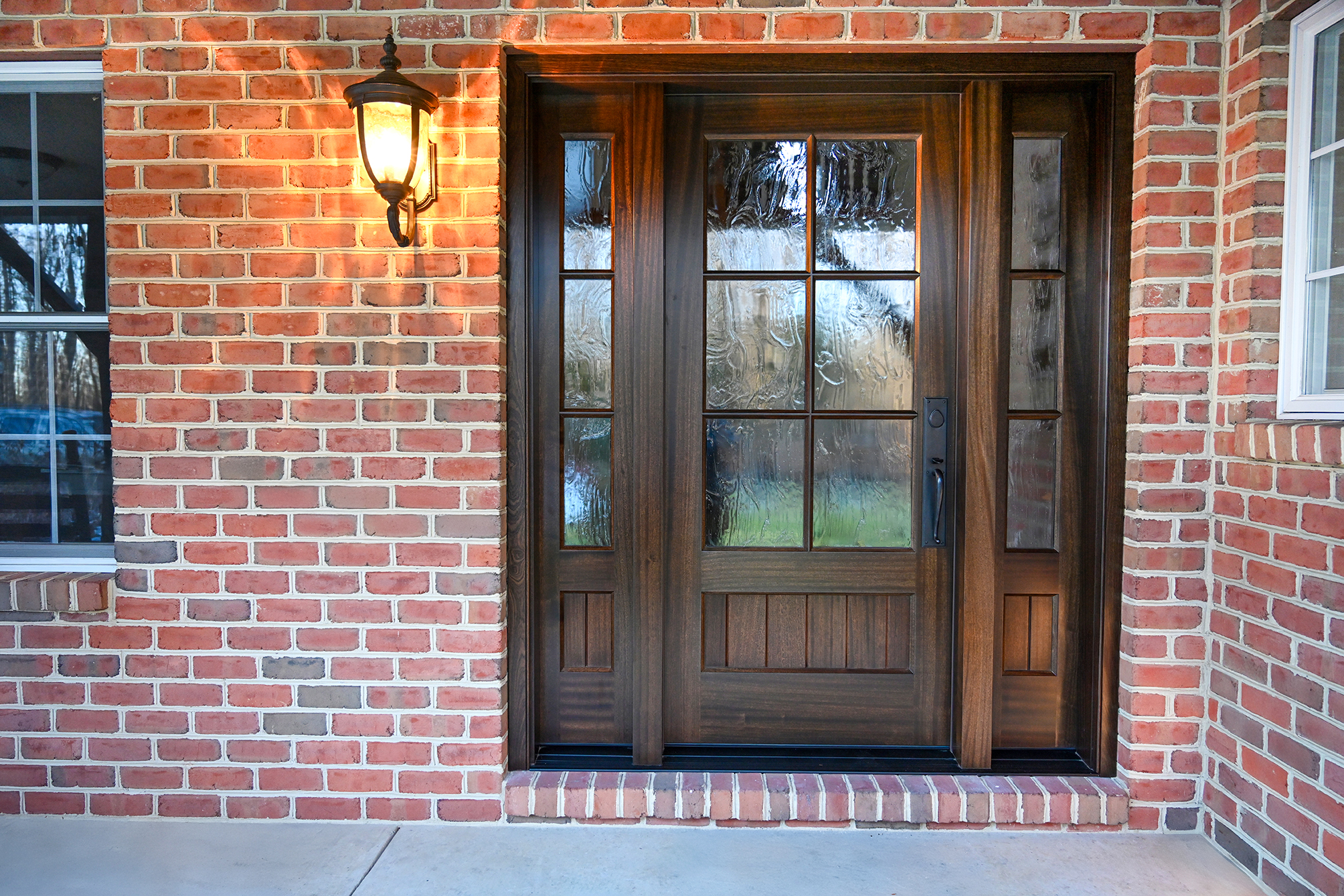 A wooden front door with glass panels is set in a red brick wall. A lit lantern-style light fixture hangs to the left of the door, and a window with white trim is partially visible. The entryway has a concrete floor.