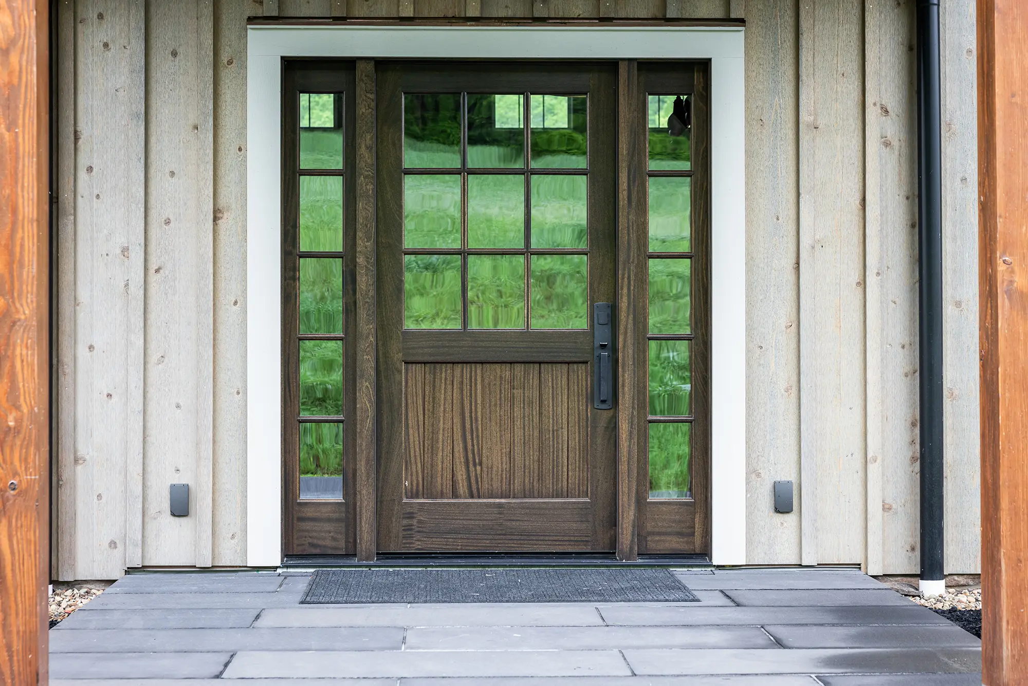 A modern wooden front door with glass panels and matching side windows, set in a light-colored wooden exterior wall. A black mat lies in front of the door on a gray tiled porch. Green trees are reflected in the glass.