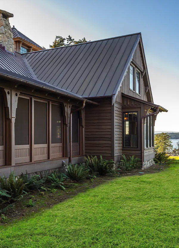 A rustic house with dark wood siding and a metal roof, featuring large windows and a screened porch, sits beside a neatly trimmed lawn with ferns, overlooking water and trees in the background.