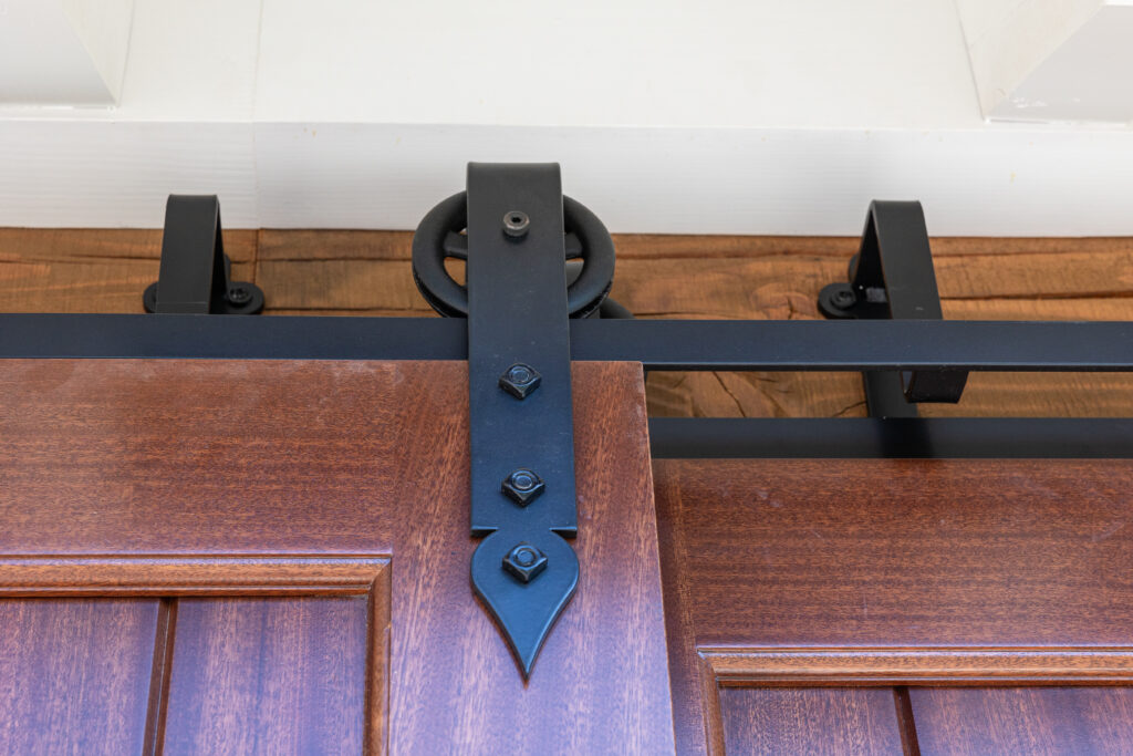 Close-up of a wooden sliding barn door with black metal hardware mounted on a wooden and white wall. The image focuses on the top part of the door and the sliding rail mechanism.
