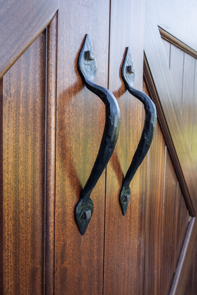 Two black metal handles with a textured finish are mounted on brown wooden double doors with a diagonal panel design. The handles are curved and positioned symmetrically, facing each other.