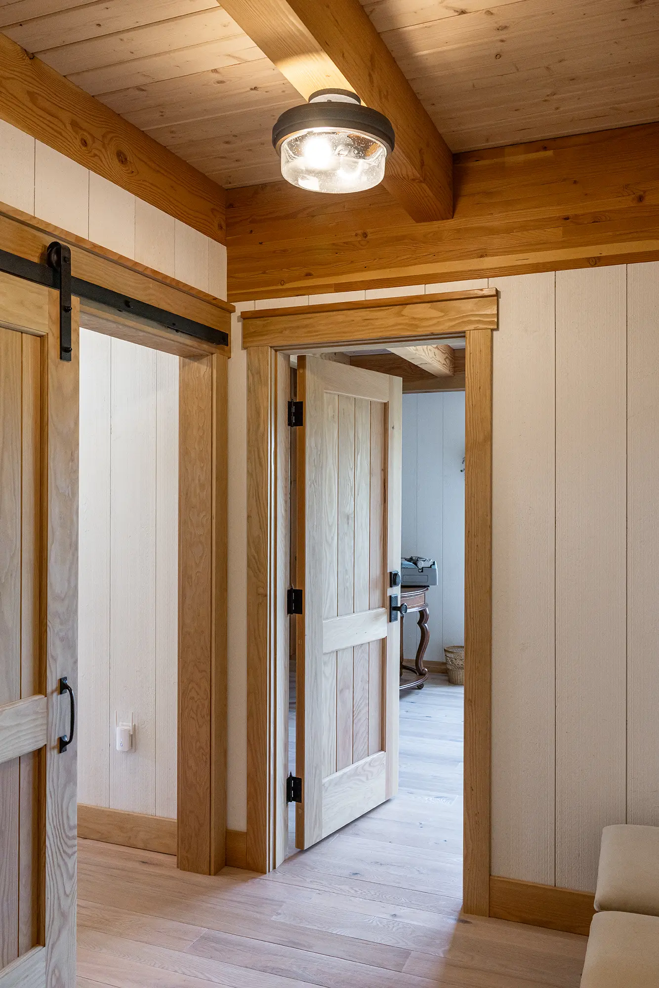A hallway in a home with light wood paneling, exposed beams, a sliding barn door, an open wooden door, and a modern ceiling light fixture. The room beyond has a wooden floor and a piece of furniture.