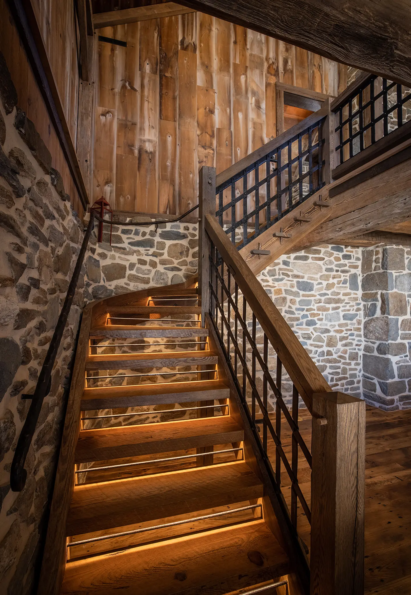 Wooden staircase with metal railings, illuminated by warm lights under each step, set against rustic stone and wood-paneled walls in a cozy, cabin-like interior.