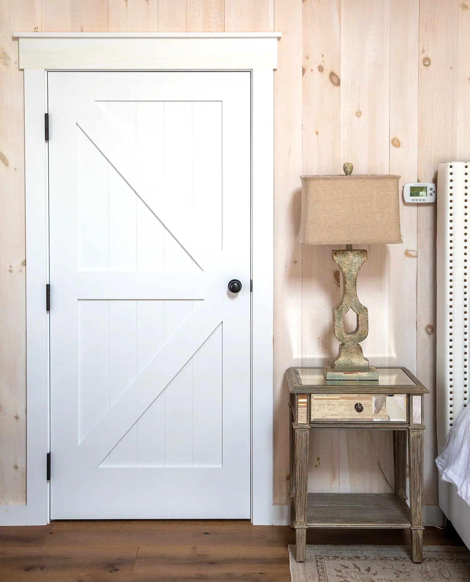 A white wooden door with a black knob is set in a light wood-paneled wall. Next to it is a mirrored side table with a beige lamp and part of a white upholstered bed visible on the right.