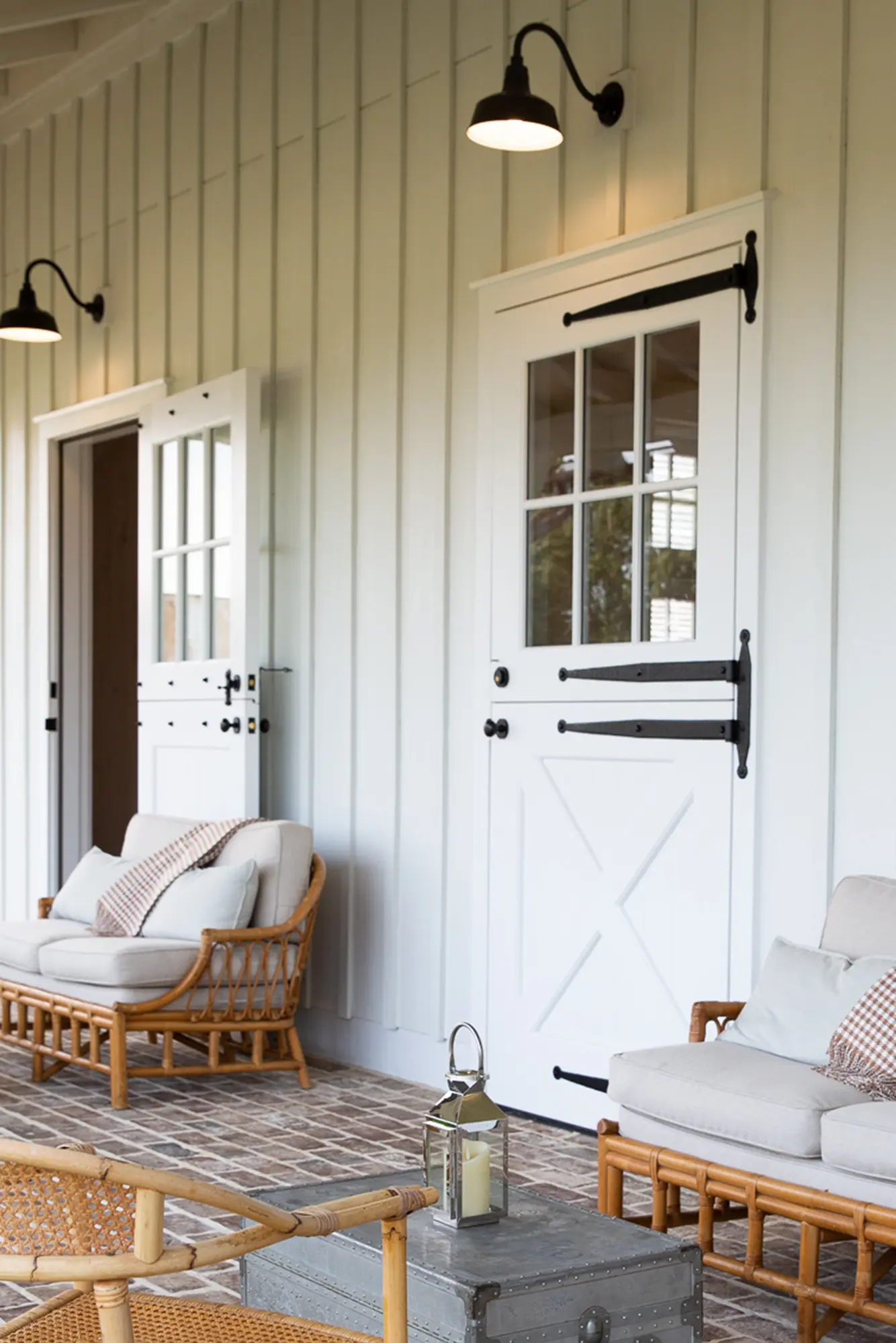 A cozy porch features two rattan armchairs with cushions, a small table, and two white Dutch doors with black hardware, all set against vertical white siding and brick flooring, lit by black wall lamps.