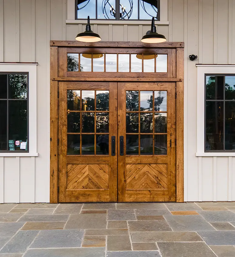 Double wooden front doors with glass panels and black handles, set in a white paneled exterior wall, flanked by two windows. Two black outdoor lights hang above the doors. Stone tiles cover the entryway.
