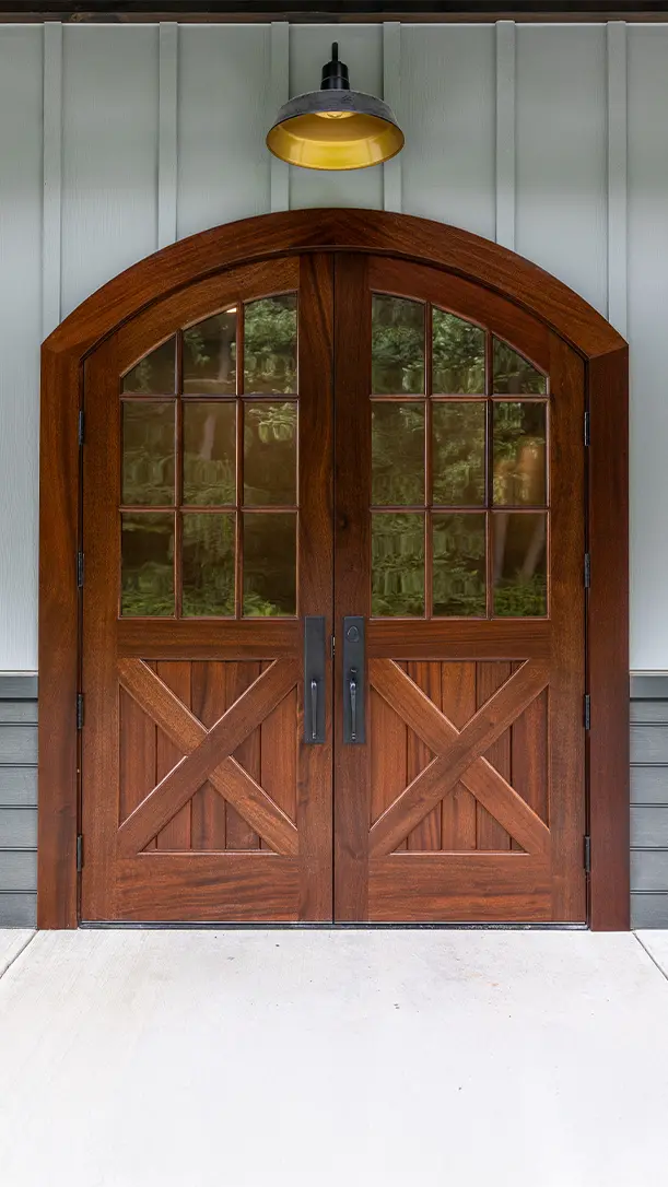 A pair of double wooden doors with an arched top, glass window panes, and decorative cross-brace panels beneath. A black metal handle is on each door, and a yellow light fixture is mounted above the entryway.