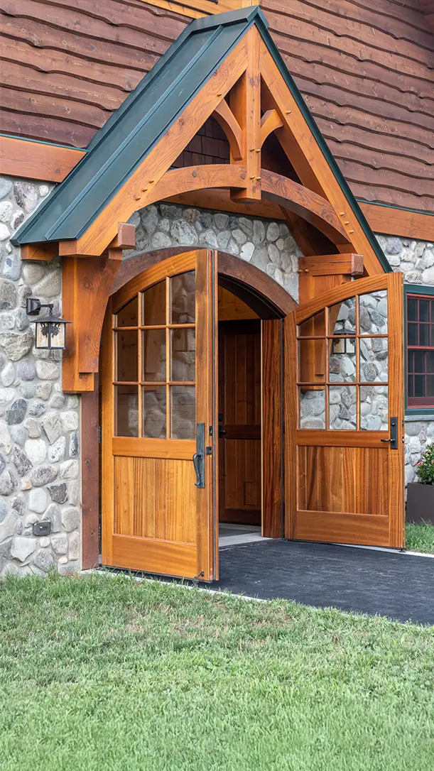 A rustic wooden double door with glass panes stands open under a peaked wooden awning, set in a stone and wood exterior wall of a building. Grass and a paved walkway are in front.