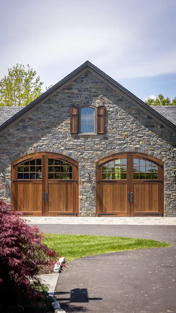 A stone garage with two large wooden arched doors and a small window with shutters above, set against a blue sky and surrounded by green grass and trees.