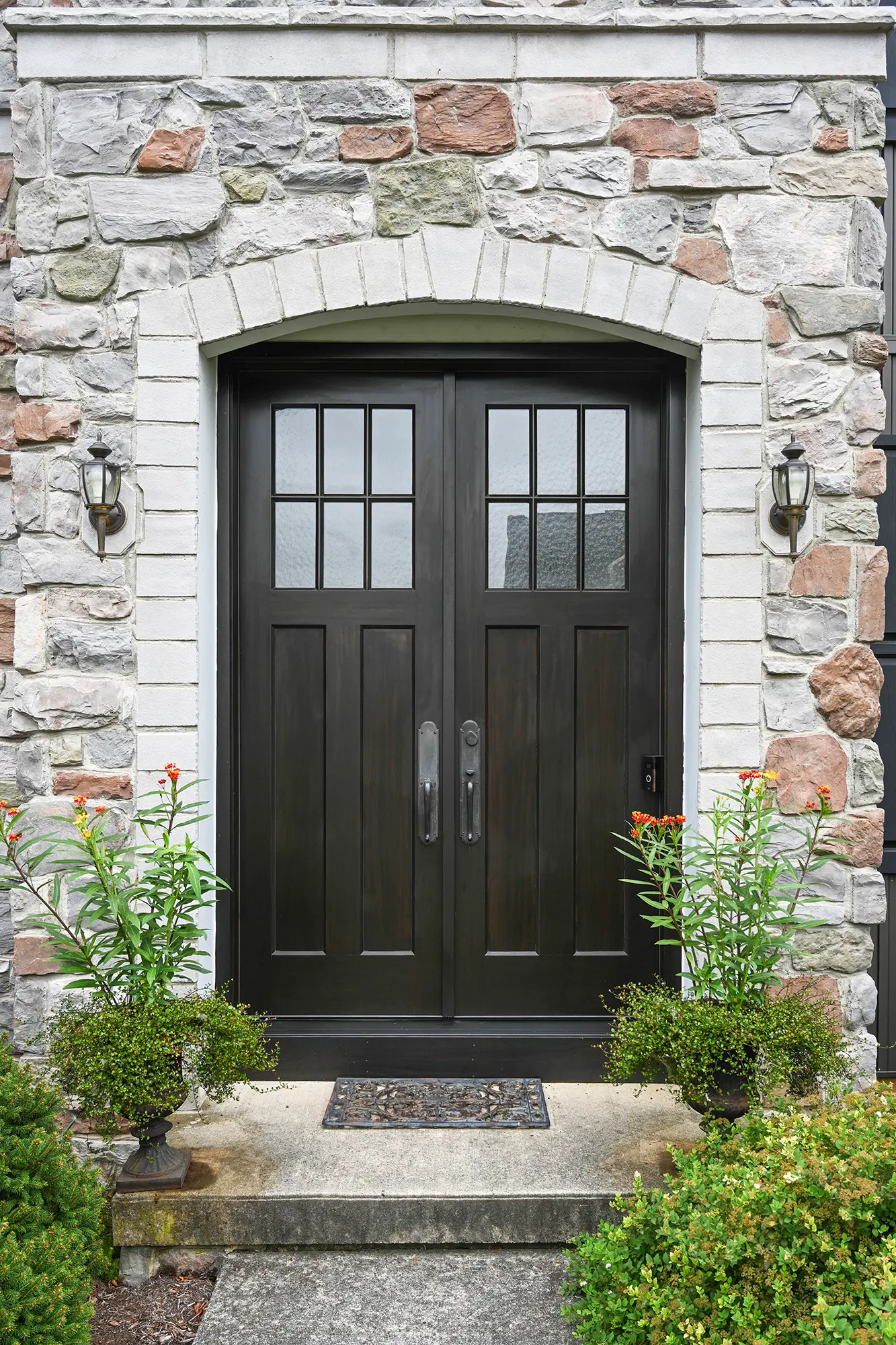A double black wooden door with glass panes is set in a stone exterior wall. Two wall lanterns and potted plants with orange flowers decorate each side of the entrance, with a small welcome mat on the concrete step.