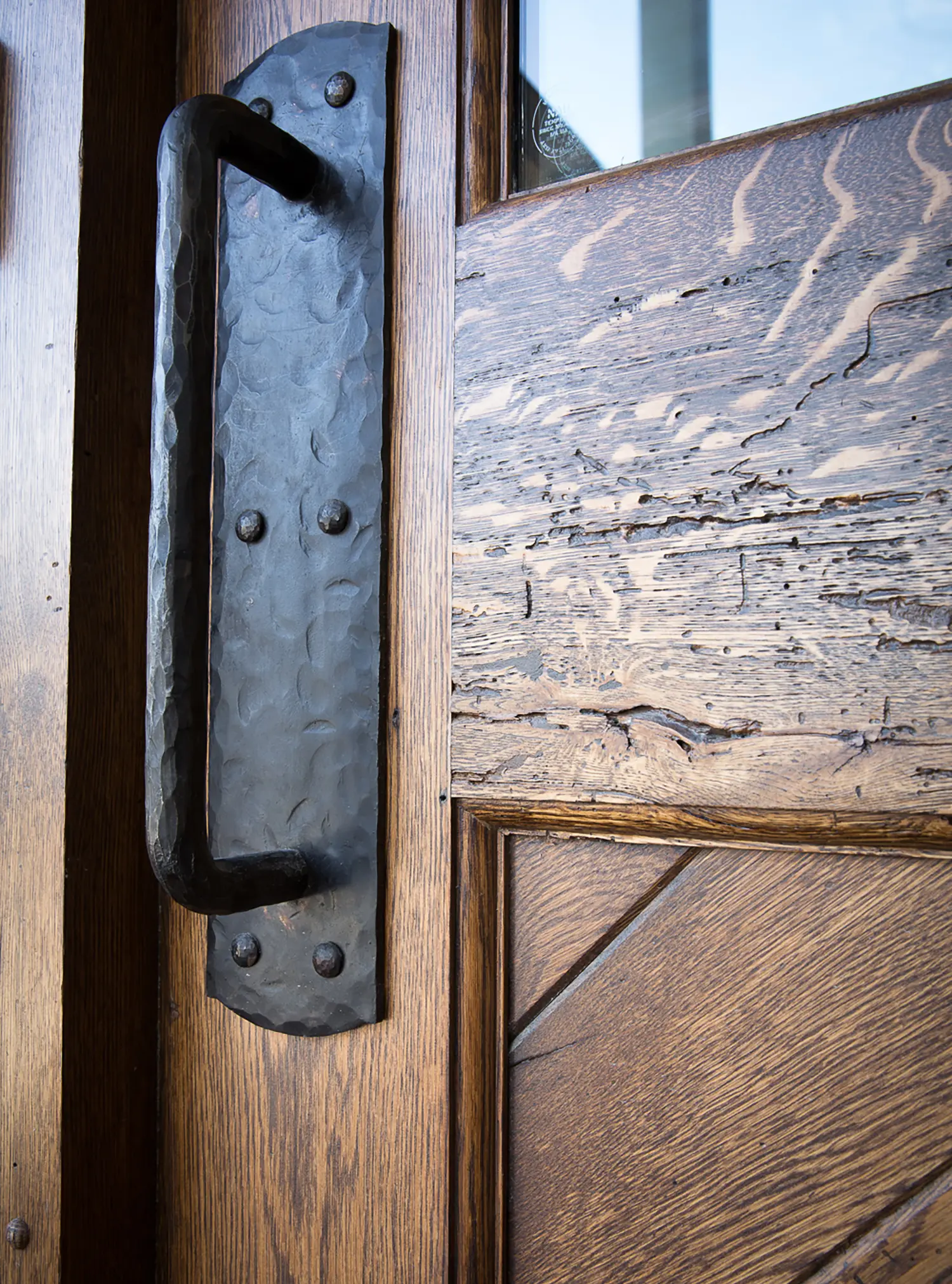 Close-up of a rustic wooden door featuring a large, textured black metal handle with visible bolts and a distressed wood finish.