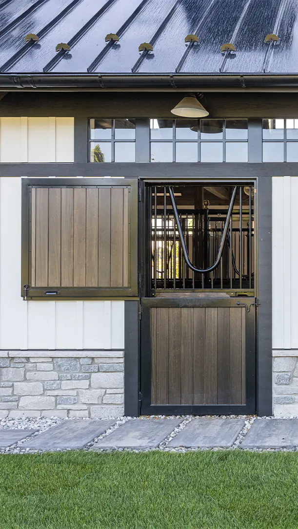 A barn door with a top half open and a bottom half closed, featuring a metal bar design. The building has white and stone walls, a black metal roof, and a path with stepping stones in front.
