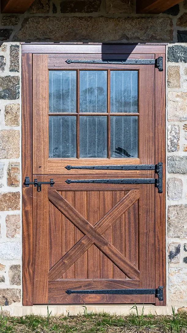 A wooden Dutch door with iron hinges and latch, featuring a window with six glass panes in the upper half, set in a stone wall.