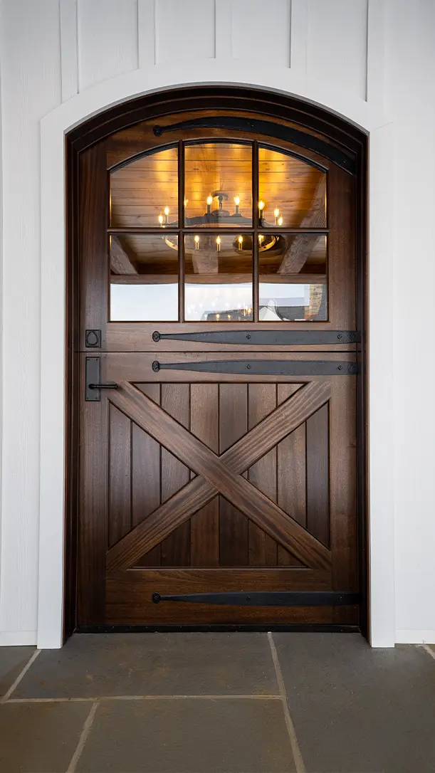 A dark wooden Dutch door with a crossbuck design and upper glass panes, set in a white paneled exterior wall. A warm chandelier is visible inside through the windows.
