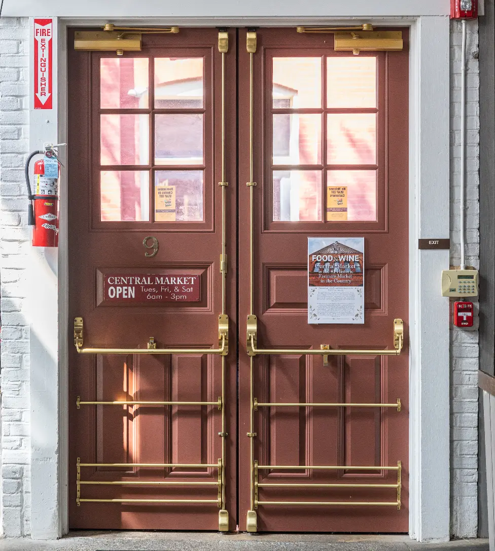 Double brown doors with brass push bars and safety features, displaying signs for Central Market hours and a food and wine event. A fire extinguisher and alarm are mounted on the white brick wall beside the doors.