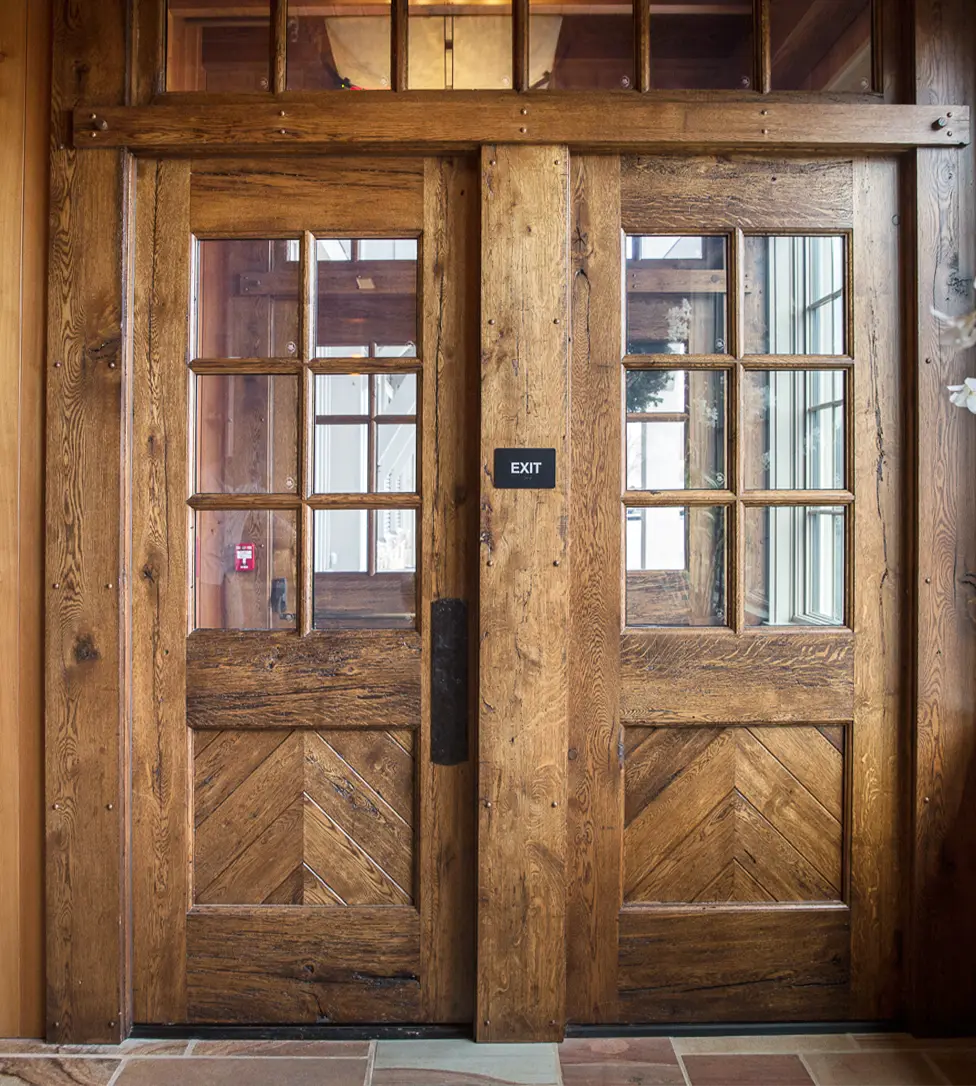 A pair of rustic wooden doors with glass panes and a chevron-patterned lower section, featuring a small black EXIT sign on the right door. Warm wooden walls and windows are visible around the doors.