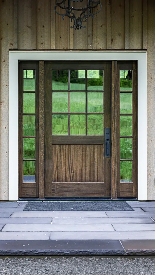 A brown wooden front door with large glass panels and sidelights, framed by white trim, set in a wood-paneled exterior wall. Gray stone tiles cover the porch floor. Green grass is visible through the glass.