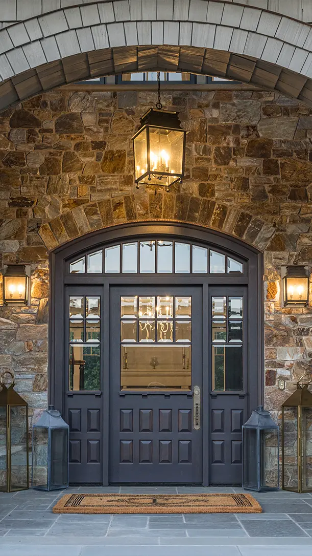 A double front door with decorative glass panels is set in a stone facade. Two lantern-style wall lights and a hanging lantern illuminate the entrance. A doormat and large lanterns sit on either side of the doorway.