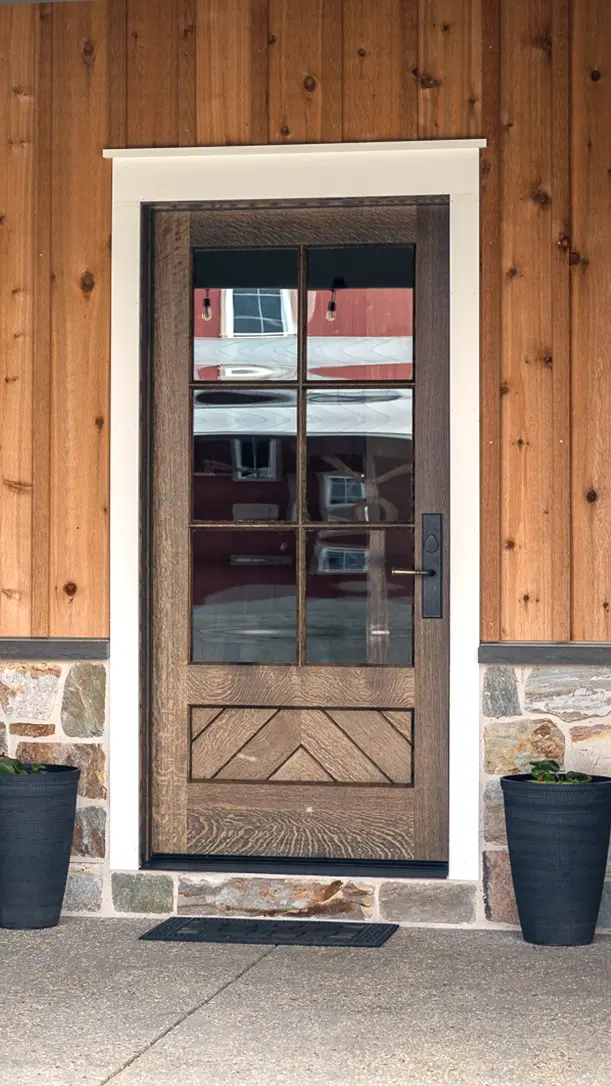A rustic wooden front door with four glass panels, white trim, and black handle, set in a wall of wood siding and stone. Two black planters sit on either side of the entrance.