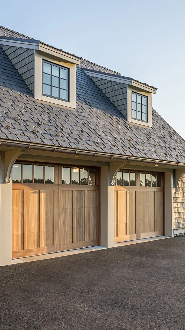 A house with two wooden garage doors, gray shingle roof, and two dormer windows, viewed in daylight from the driveway.