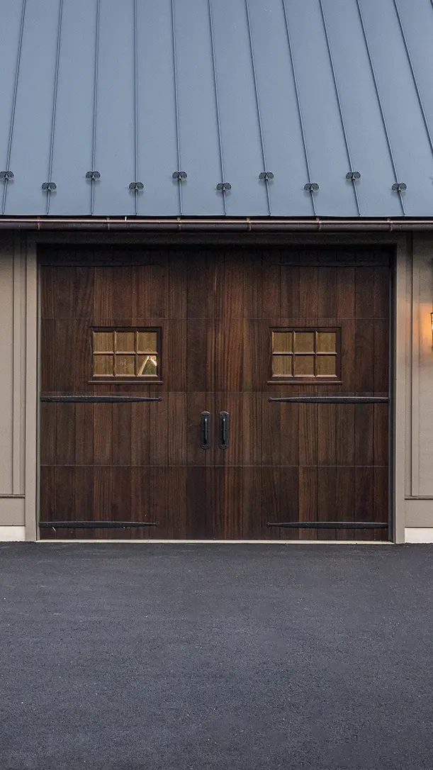A modern building with a dark wooden double garage door featuring two small square windows, set below a metal roof and surrounded by gray siding, with an asphalt driveway in front.