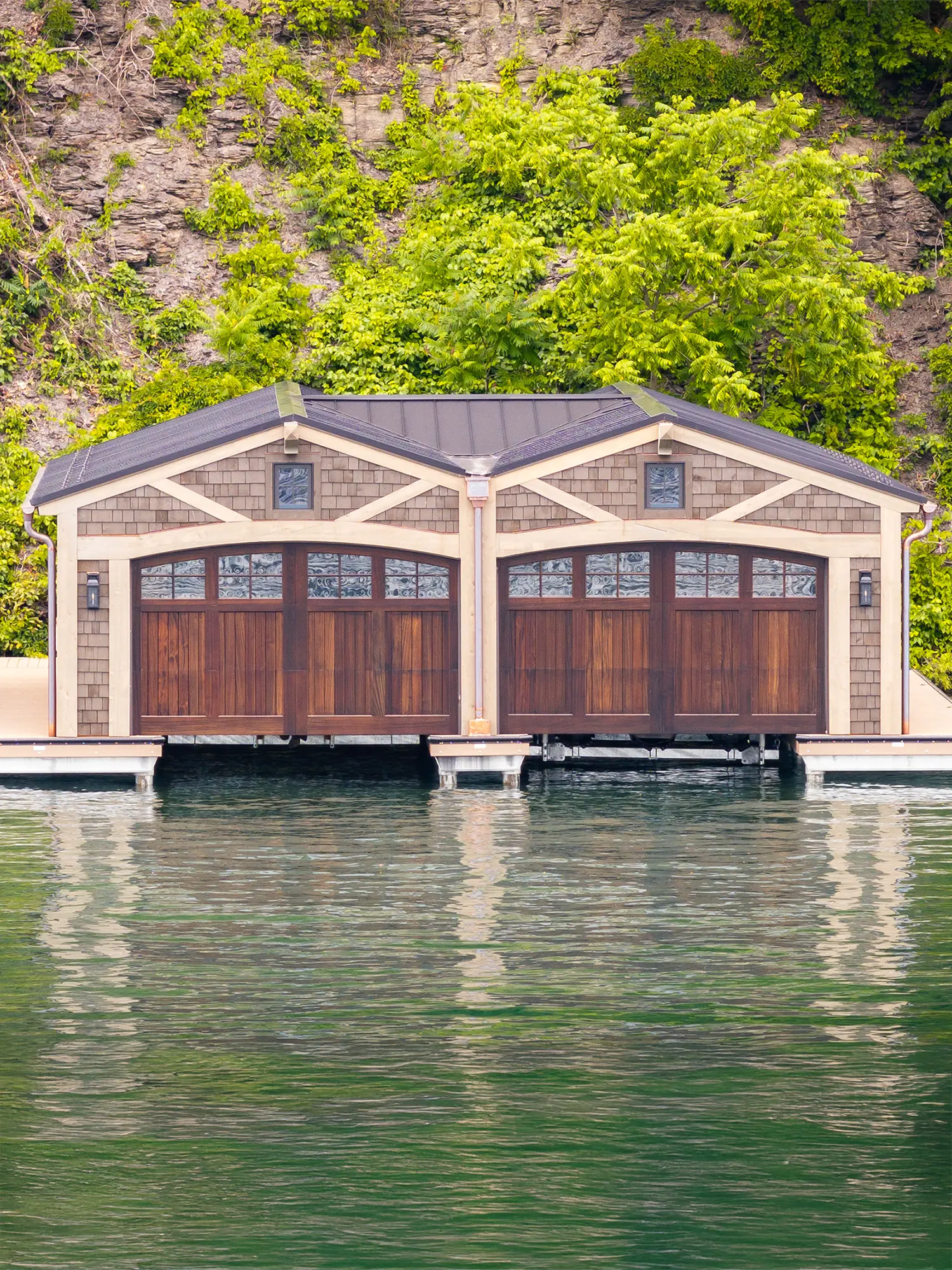A boathouse with two wooden garage doors sits on the edge of calm water, backed by a rocky cliff covered in green foliage.