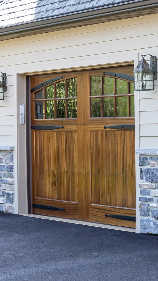 Wooden garage door with decorative black metal hinges and handles, small windows at the top, and stone accents on the exterior wall. Two lantern-style light fixtures are mounted on either side of the door.