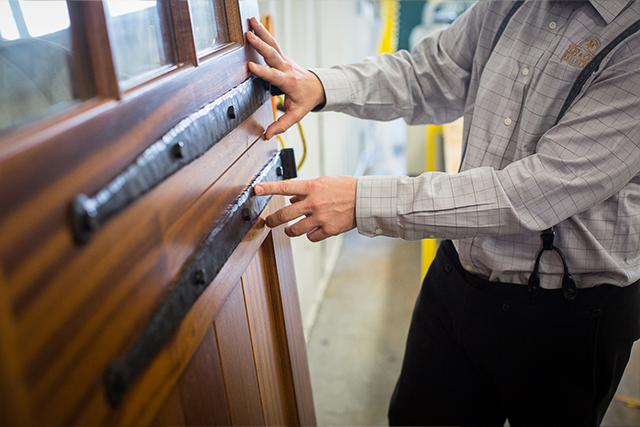 A person wearing a plaid shirt and suspenders inspects or installs a wooden door, touching its decorative metalwork strip with both hands.