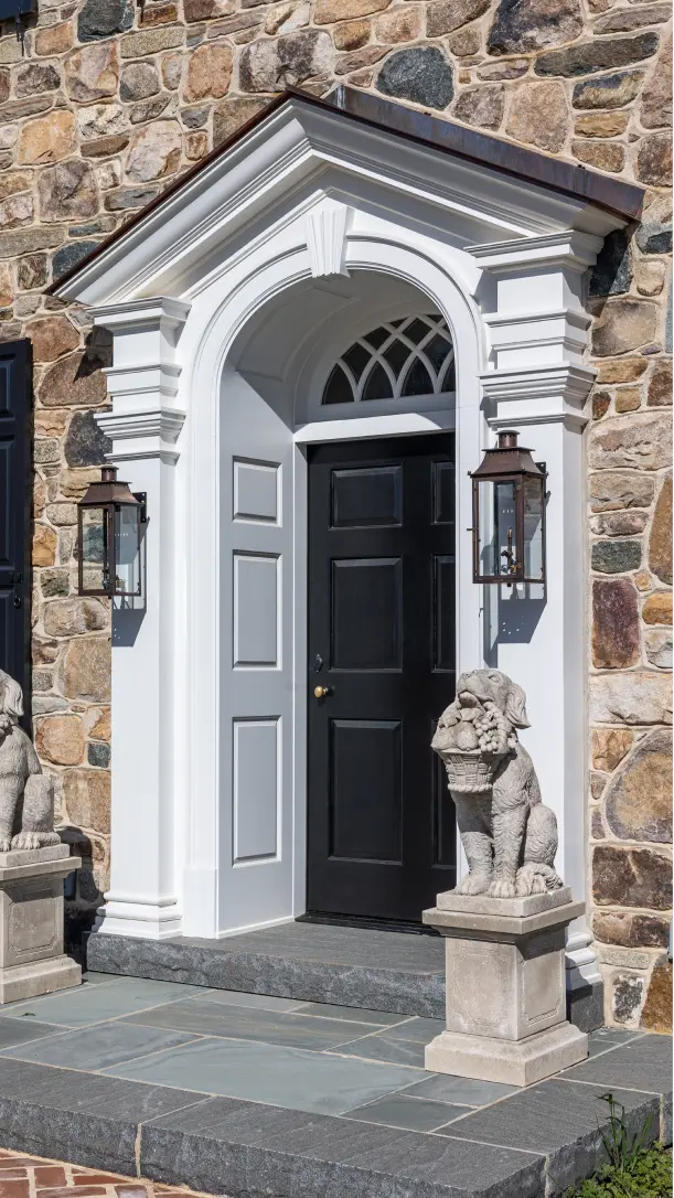 A black front door with a white decorative archway, flanked by two stone statues of bulldogs on pedestals, and two lantern-style wall lights, set in a stone exterior wall.