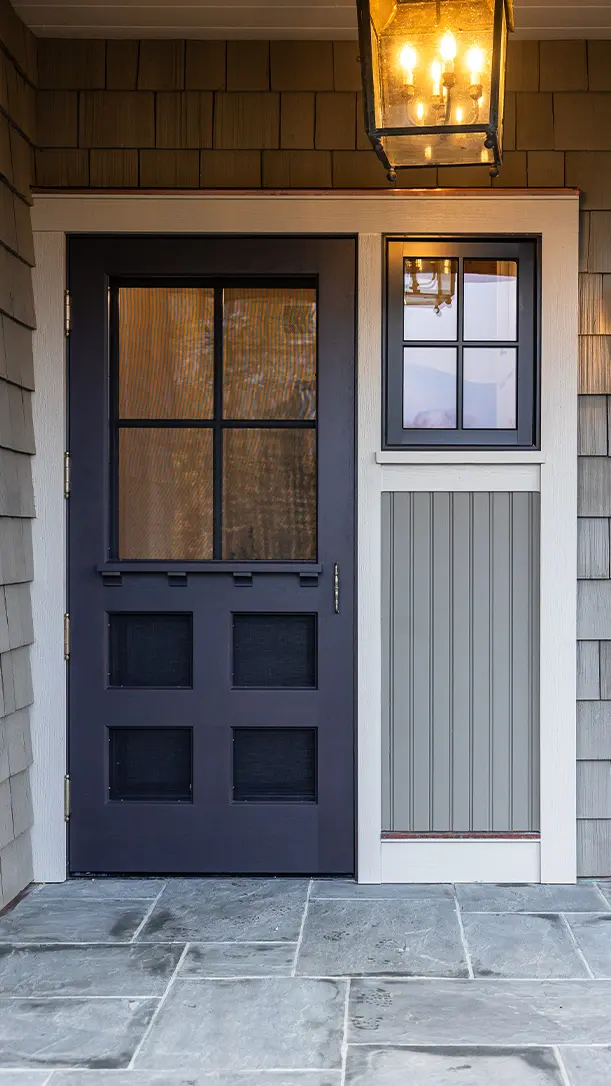 A modern front door with four glass panes and a side window, framed in white trim, set against gray shingled siding and gray stone tile flooring, with a black hanging lantern overhead.