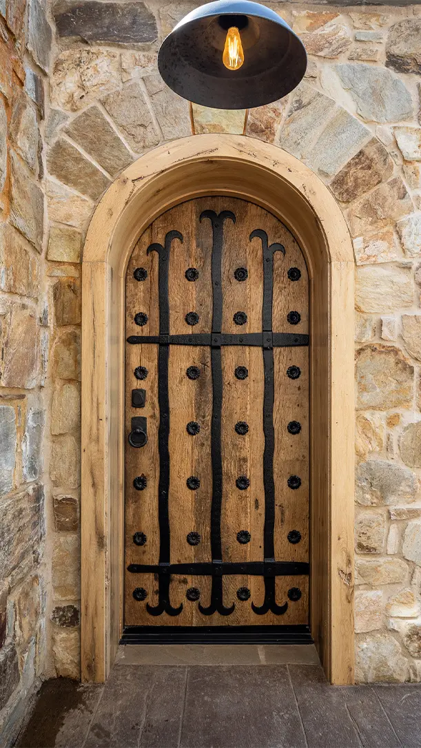 A rustic arched wooden door with decorative black ironwork and studs is set in a stone wall, with a single exposed bulb pendant light hanging above the entrance.