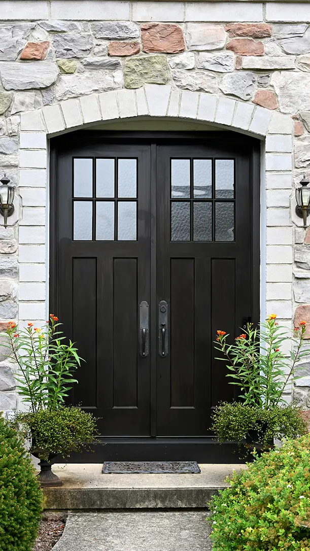 A pair of black double doors with glass panels at the top set in a stone exterior wall, flanked by two potted plants with orange flowers and small outdoor wall lights on either side.