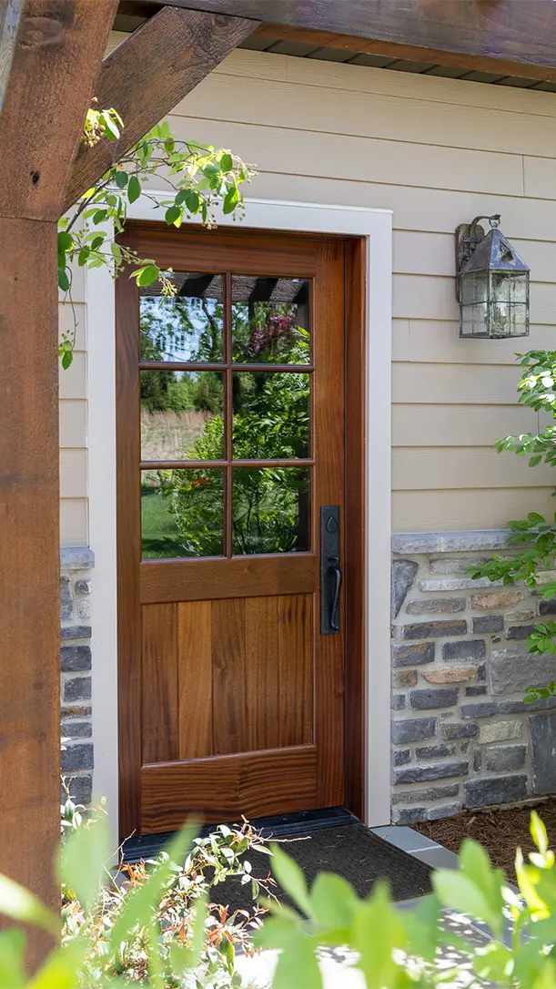 A wooden front door with a windowpane design and black handle, set in a stone and beige siding exterior wall. Green plants and a rustic outdoor lantern are nearby.