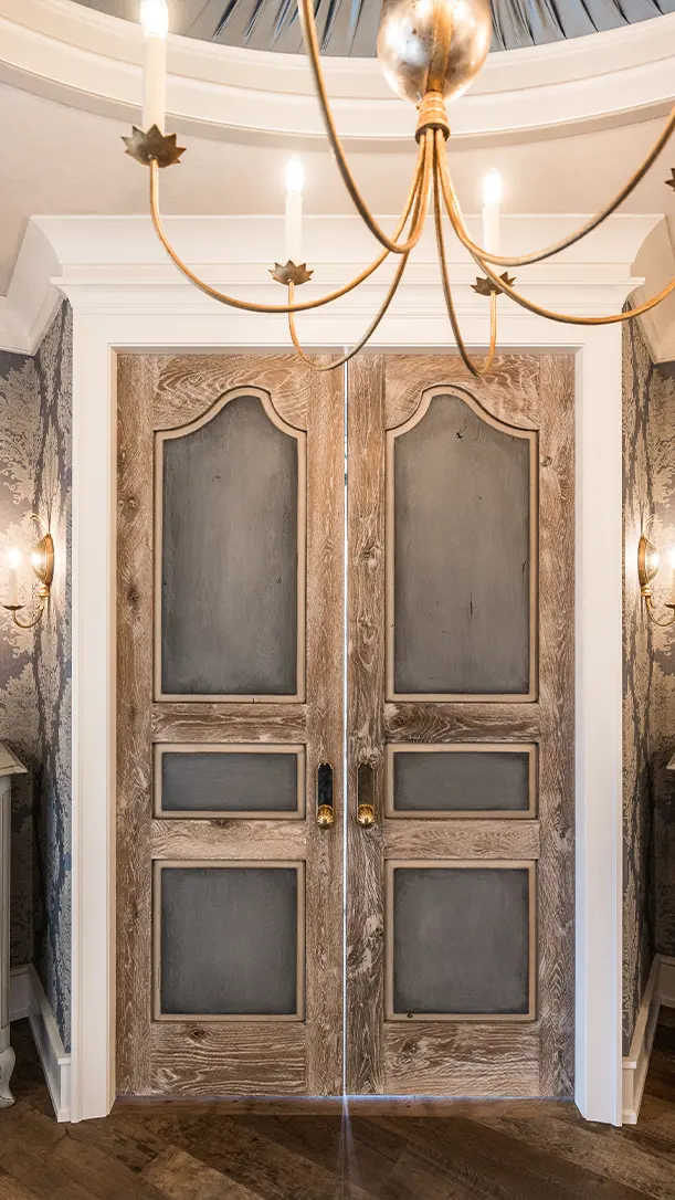 Double wooden doors with dark gray panels and ornate trim, framed by white molding. A brass chandelier with exposed bulbs hangs above, and patterned wallpaper decorates the surrounding walls.