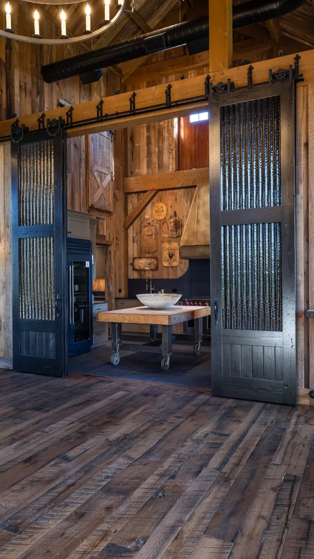A rustic kitchen with wooden floors and walls, featuring a central island with a sink on wheels, black sliding barn doors with glass panels, and various kitchen tools hanging on the back wall.