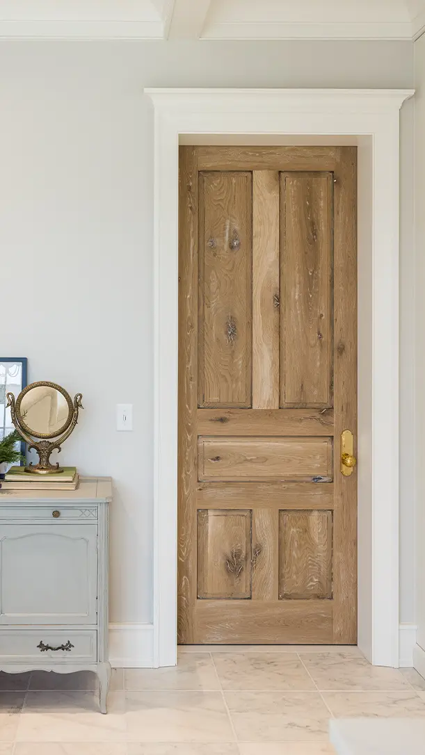 A wooden door with a natural finish and gold handle is set in a white-trimmed doorway. Next to it is a light gray vintage-style dresser with a round mirror and decorative items on top.