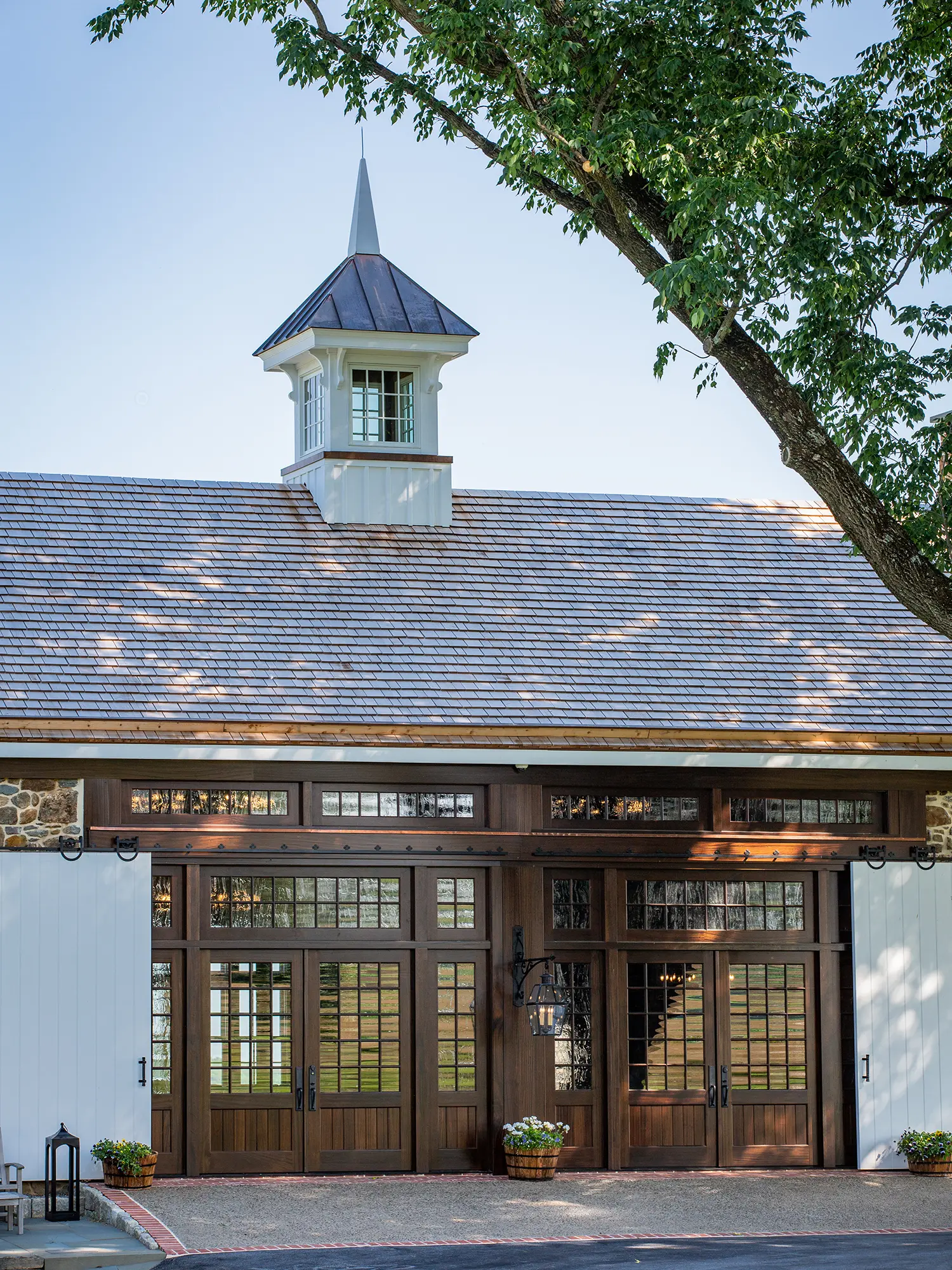 A building with a wooden exterior, large glass-paneled double doors, a cupola on the roof, and potted plants by the entrance, shaded by a leafy tree in the foreground.