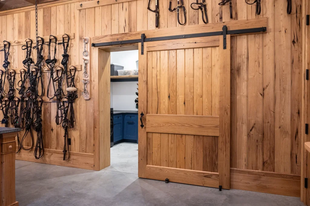 A sliding wooden barn door opens to reveal a room with blue cabinets. The wood-paneled walls have hooks holding horse bridles and tack. The floor is smooth concrete.