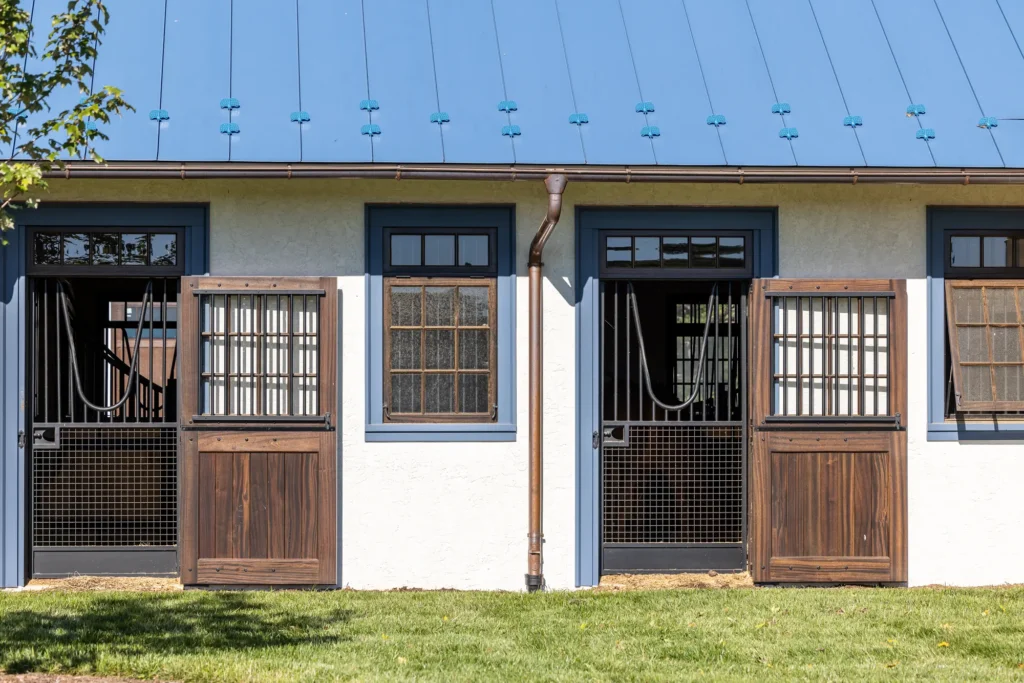 Exterior view of a stable with three stalls, each featuring wooden half-doors and metal bars, under a blue metal roof, with grass and part of a tree visible in the foreground.