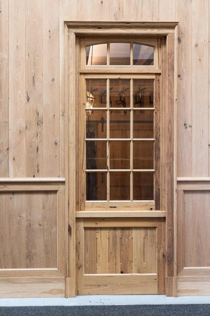 A wooden door with glass window panes set in a light wood paneled wall, with a glimpse of an interior lantern and wooden details visible through the glass.