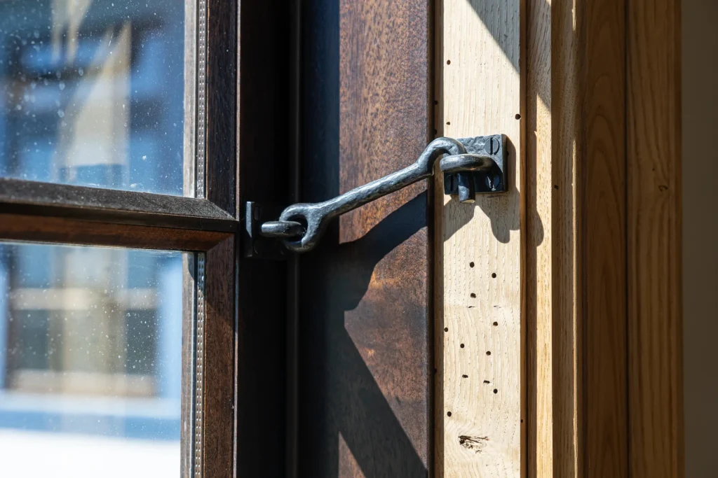 Close-up of a wooden window secured with a metal hook latch in bright sunlight, showing part of the frame, latch, and a slightly open glass window.