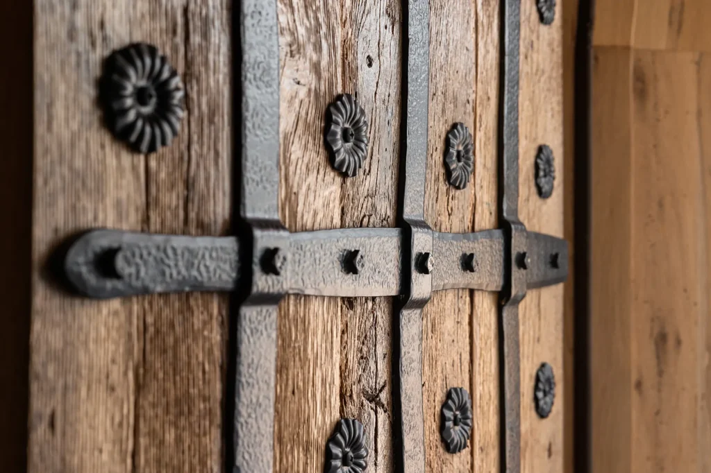 Close-up of a rustic wooden door with black wrought iron hinges and decorative rosettes, showing the texture of the aged wood and metal details.