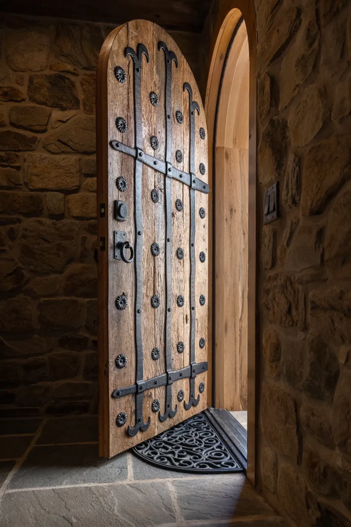 A heavy wooden door with ornate black iron hinges and studs stands open, revealing part of a stone wall and a dark stone floor. Light spills in through the doorway, highlighting the textures of the wood and stone.