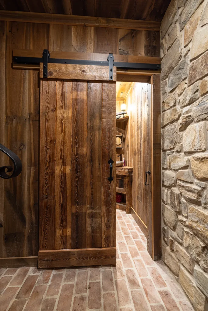 A rustic wooden sliding barn door is partially open, revealing a warmly lit room with wooden walls and shelves. The floor is made of red bricks, and one wall is constructed of large, irregular stones.