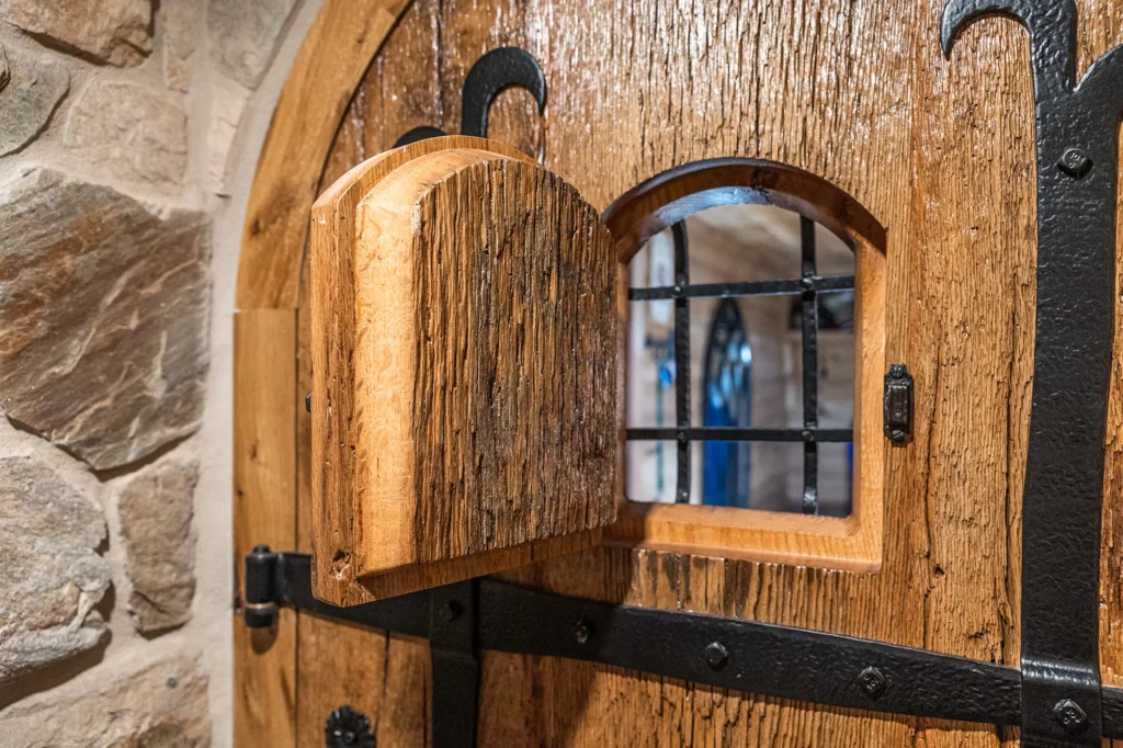 A close-up of a rustic wooden door with a small hinged hatch open, revealing a window with black metal bars. The door has iron hardware and is set in a stone wall.