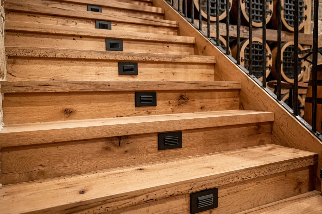 Close-up view of wooden stairs with built-in rectangular lights on each step, black metal railings on the sides, and large wine barrels visible in the background.