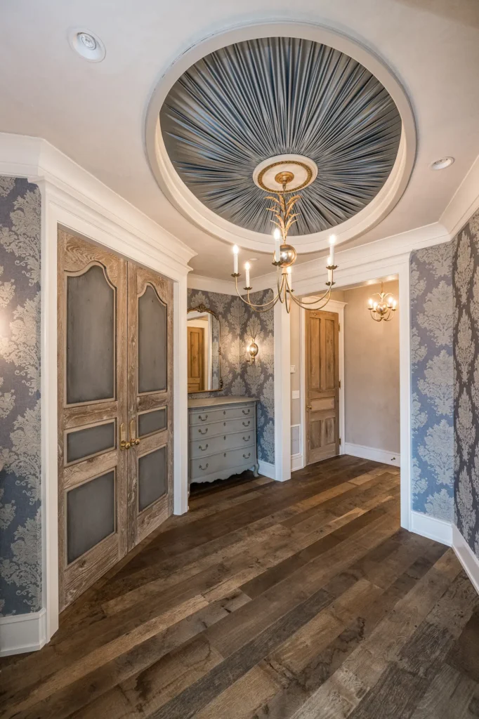 Elegant hallway with dark wood floors, ornate double doors, vintage dresser, floral-patterned wallpaper, and a ceiling with a blue sunburst design and gold chandelier.