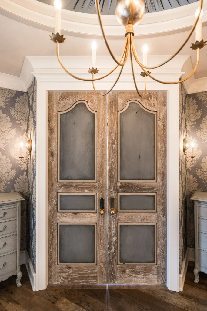 Double wooden doors with ornate panels, set in a white frame, below a brass chandelier. The room has patterned wallpaper, wall sconces, and two light gray dressers on either side of the doors.